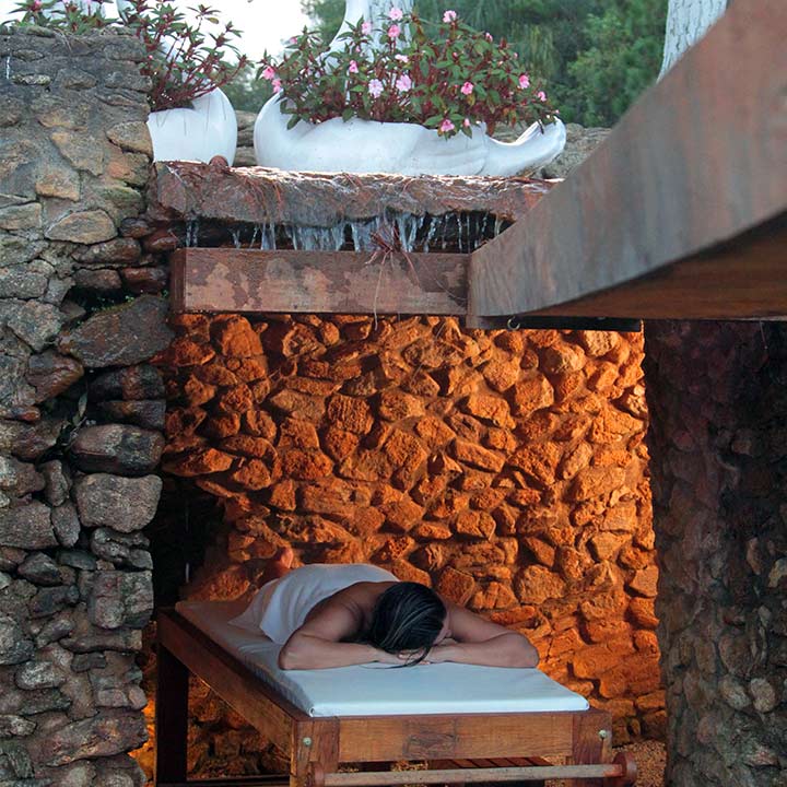 Pessoa deitada relaxando em um spa ao ar livre com estrutura de pedra e iluminação aconchegante, cercada por plantas e flores em vasos brancos.