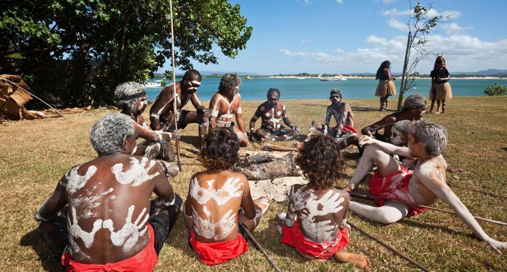 A photo showing the Guugu Yimithirr people of Australia sitting in circle at the beach with relaxed expressions.