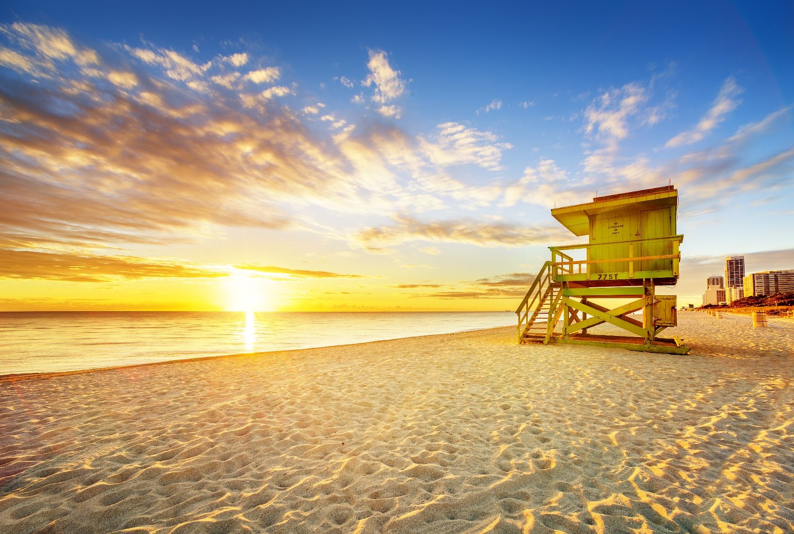 Sunrise at South Beach in Miami, Florida, showing a green lifeguard tower on the sandy beach with golden light reflecting off the ocean and clouds in a vibrant morning sky.