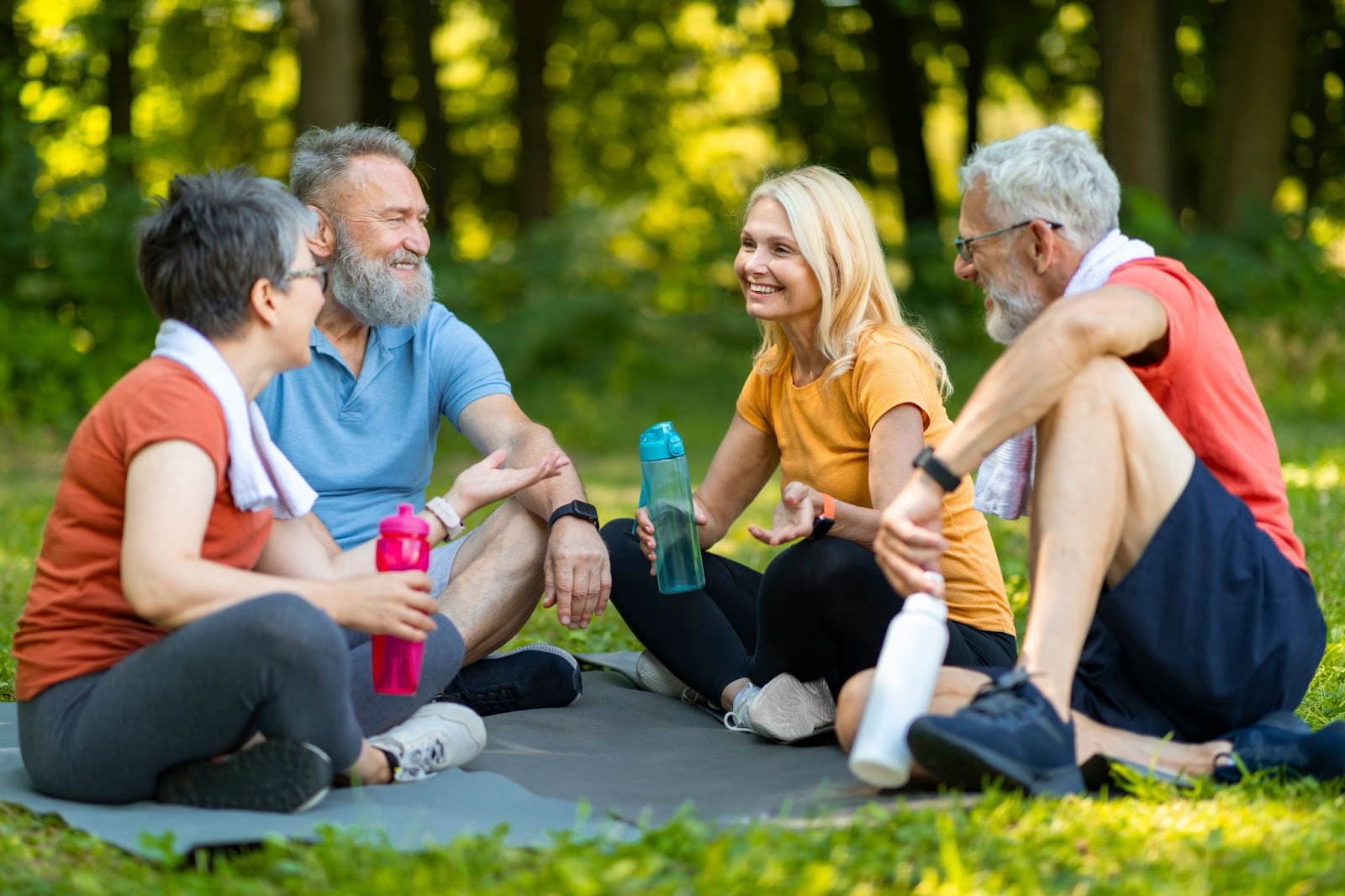 4 seniors sitting outside on yoga mats and talking while holding water bottles.