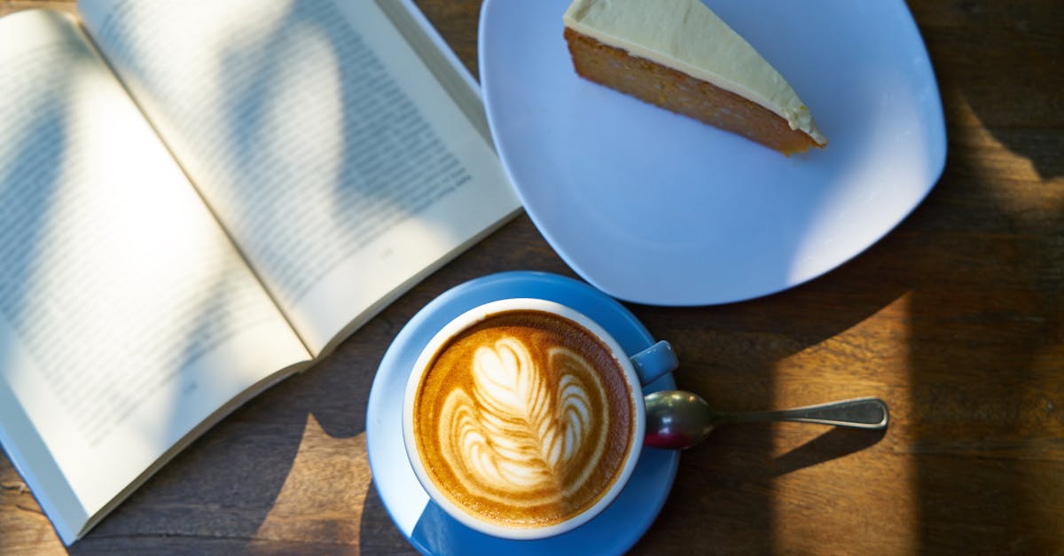 An open book with white pages lies on a wooden table bathed in sunlight. Beside it, a blue cup holds a steaming cappuccino adorned with a heart-shaped design, and a silver spoon rests on the saucer. A white plate displays a slice of cake with a white topping. The overall image evokes a sense of tranquility and leisurely enjoyment.