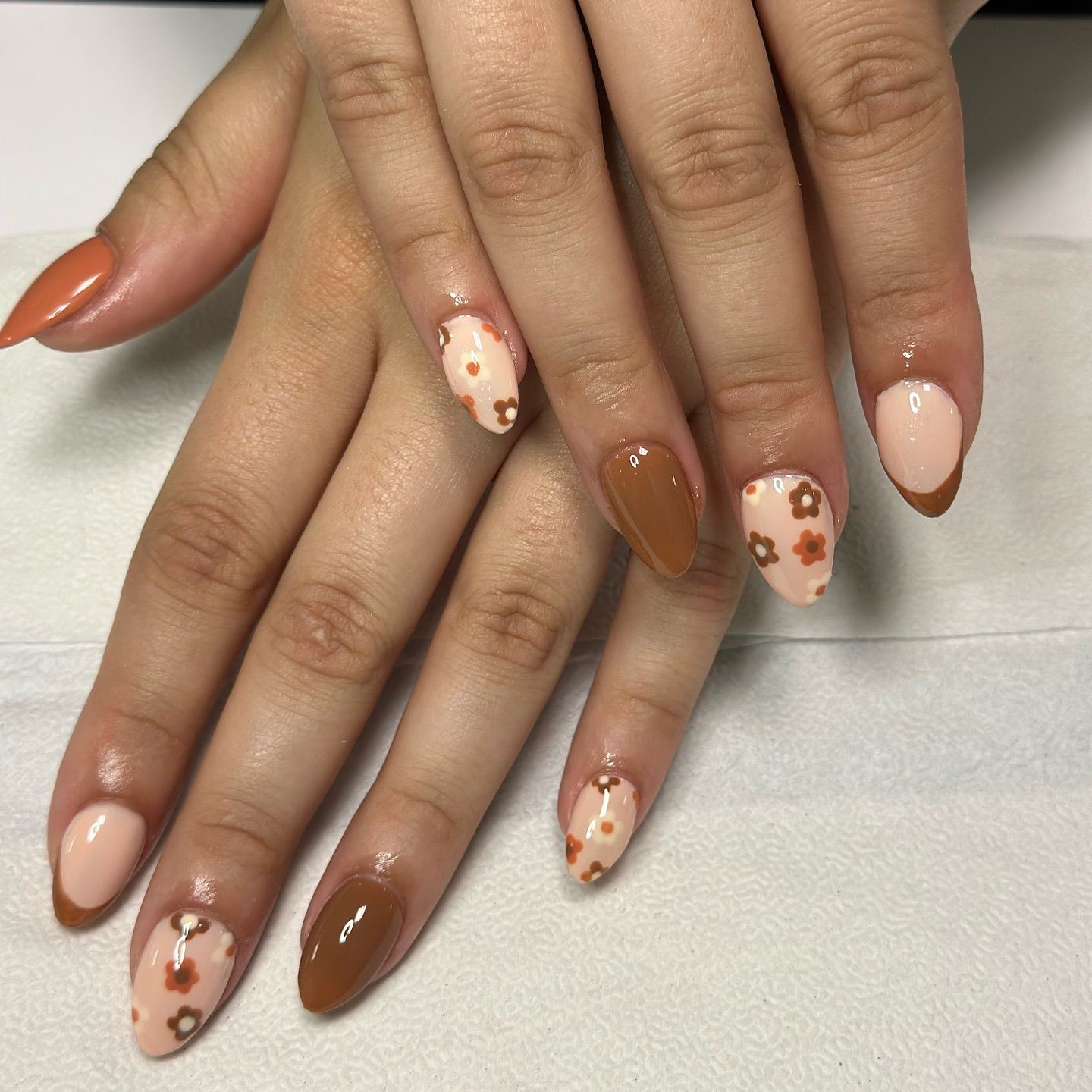 Two hands with almond-shaped nails, featuring a glossy nude polish and brown floral accents.

