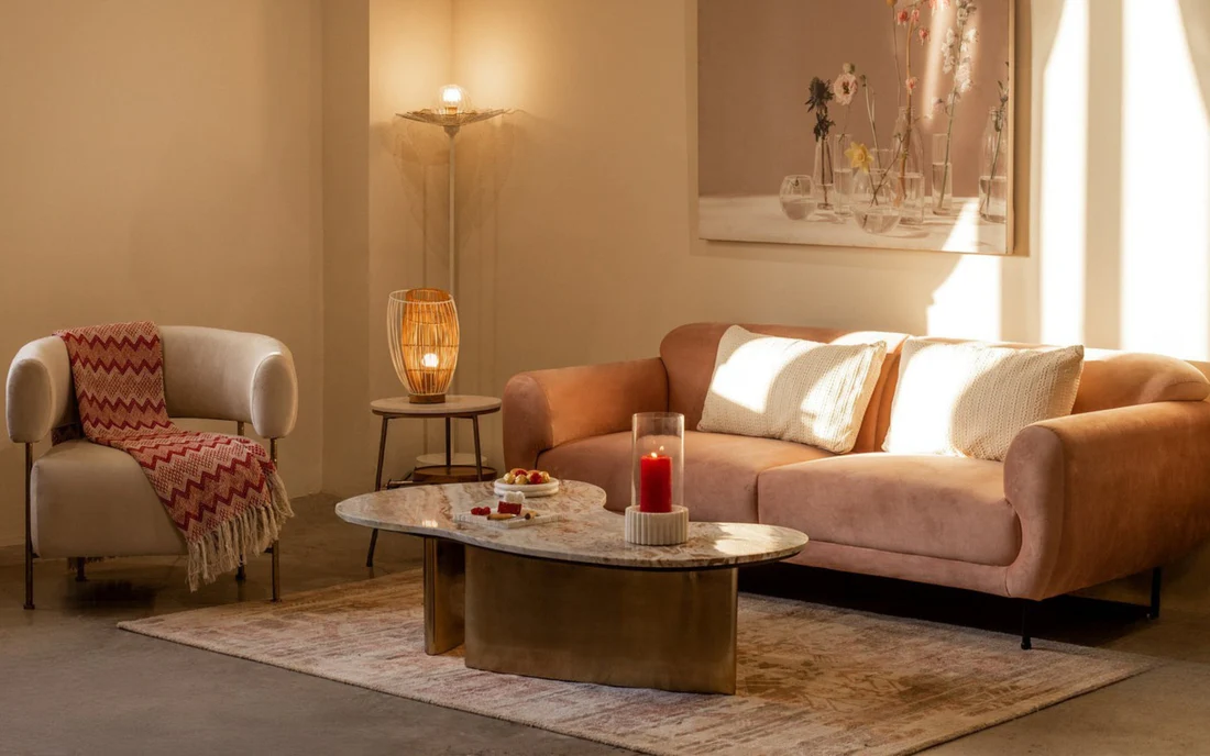 Cozy living room with a blush pink velvet sofa, cream armchair with a red throw, marble-top coffee table, warm ambient lighting from the floor and table lamps