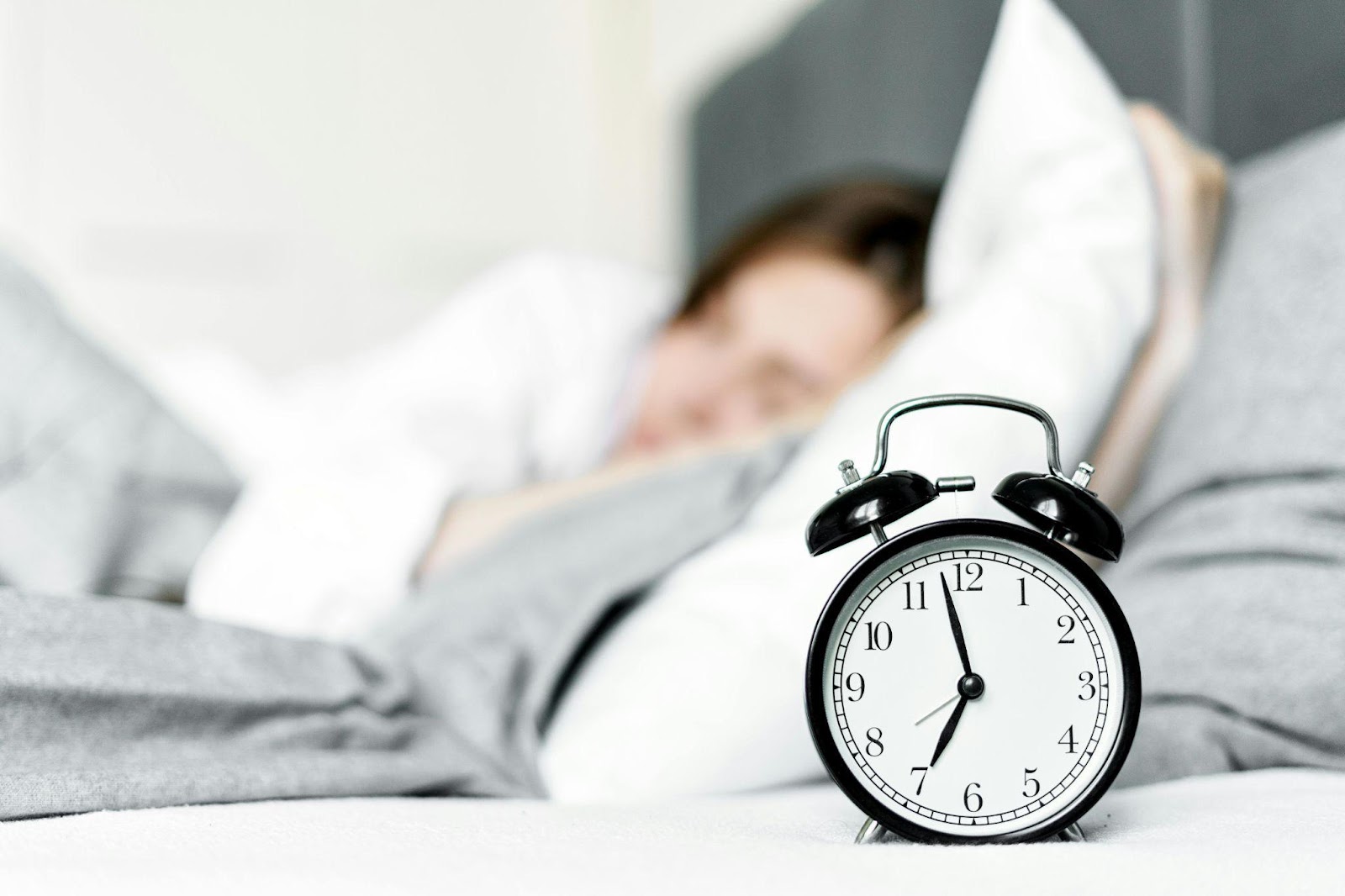 an old alarm clock reading seven o’ clock in the foreground, with a blurred woman dozing in the background