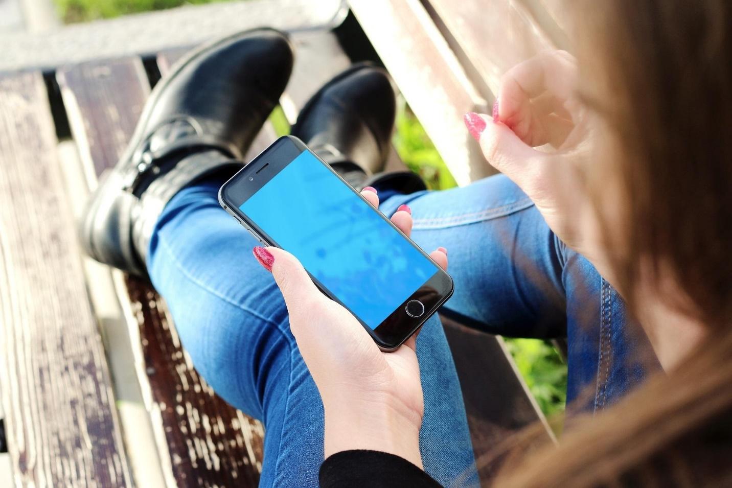 A woman sitting on a bench outdoors, holding a smartphone with a blank blue screen, wearing blue jeans, black boots, and red nail polish, representing mobile learning or digital accessibility.