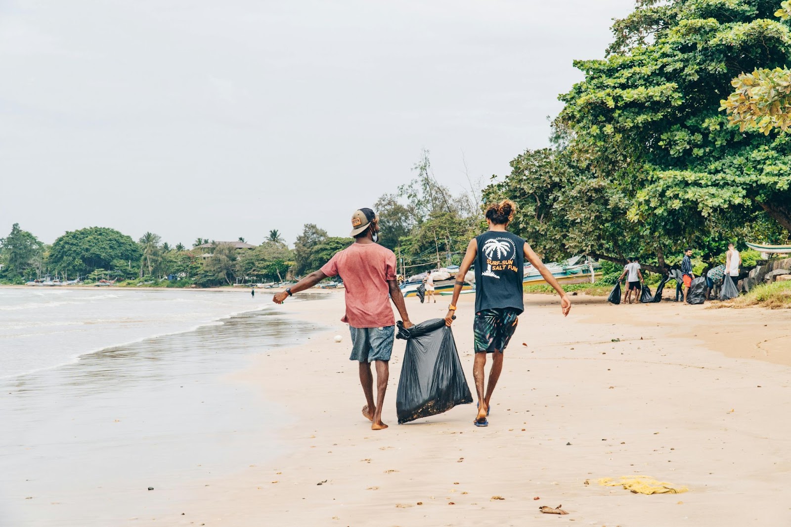 Foto de pessoas fazendo ação de limpeza na praia, com foco em dois homens carregando um saco de lixo cheio.