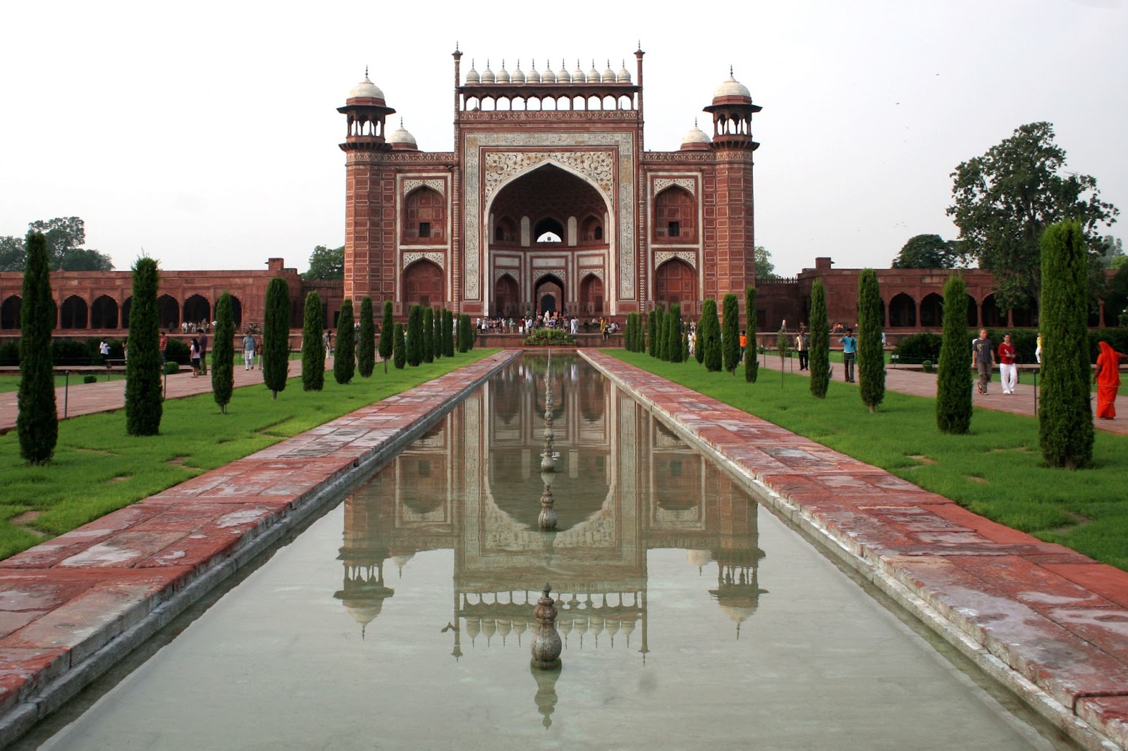 A symmetrical view of the main entrance gate to the Taj Mahal, an architectural masterpiece in Agra, features a reflecting pool and lush greenery. The red sandstone structure is adorned with intricate white marble inlays and topped with small domes.