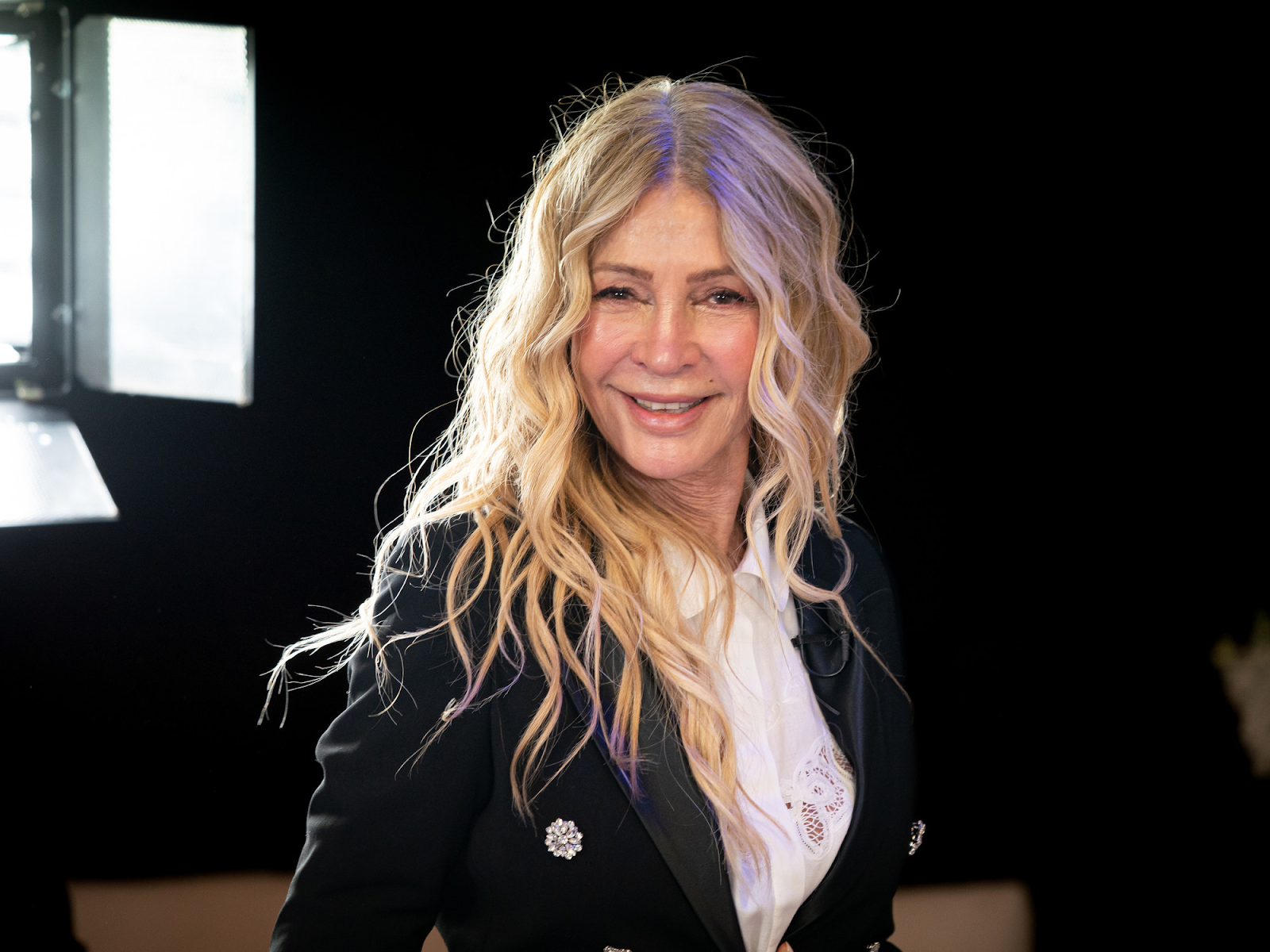 Mujer con cabello rubio rizado y chaqueta negra sonriendo en un estudio con iluminación profesional.