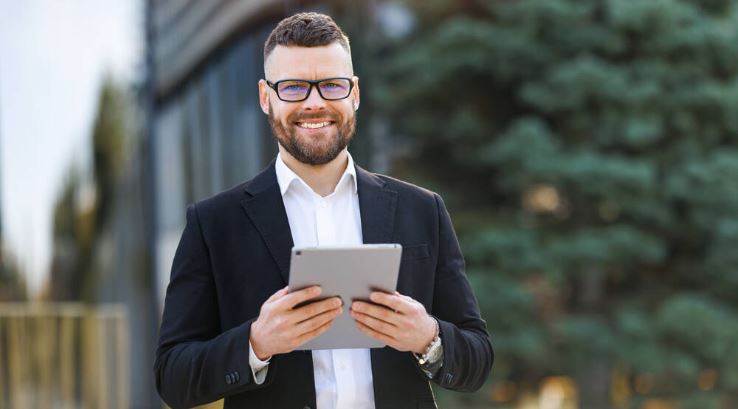 Young cheerful business man looking at digital tablet with smile while standing on sunny street outdoors