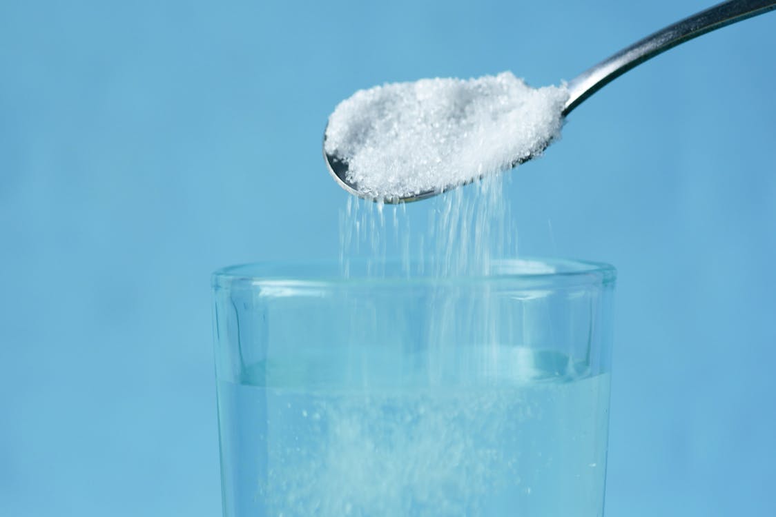 Free Pouring sugar into a glass of water against a blue background. Stock Photo