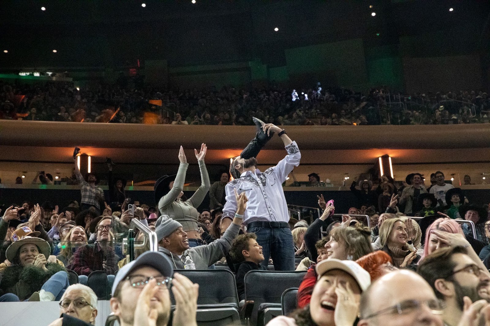 Audience member in a blue shirt drinks beer out of black cowboy boot amid cheering crowd.