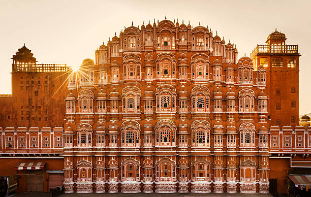 hawa mahal jaipur ,The image shows a vibrant depiction of Hawa Mahal, Jaipur, highlighting its stunning pink sandstone facade with intricate latticework and arched windows. The bright daylight and clear blue sky emphasize the architectural elegance of this historical monument, complemented by greenery and a few visitors in the foreground.