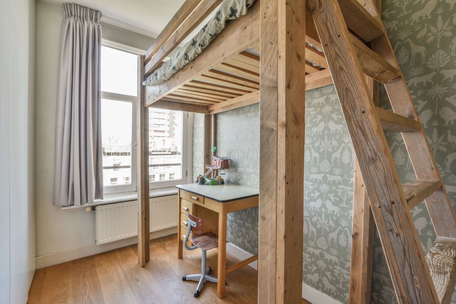 A charming lofted bed in light brown wood featuring a cozy workspace underneath, set against pastel green and white wallpaper for a contrasting, serene look.