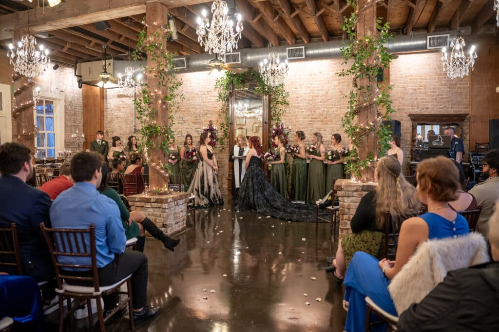 Two brides exchange vows in the elegant Grand Hall at Butler's Courtyard, an LGBTQ friendly wedding venue near Houston, Texas. Guests comfortably witness the indoor ceremony, showcasing the venue's charm and suitability as a perfect wedding location, ideal for avoiding heat or as a dependable Plan B.