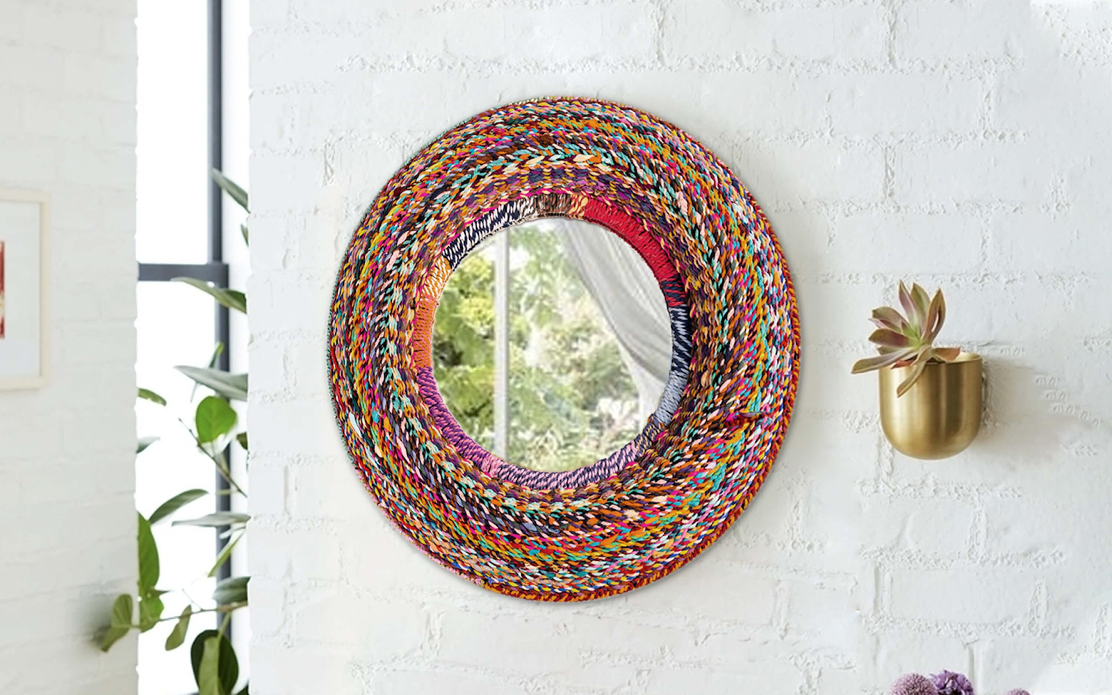 Round mirror with a vibrant, multicolored braided frame hanging on a white brick wall, next to a brass wall-mounted planter with a small succulent.