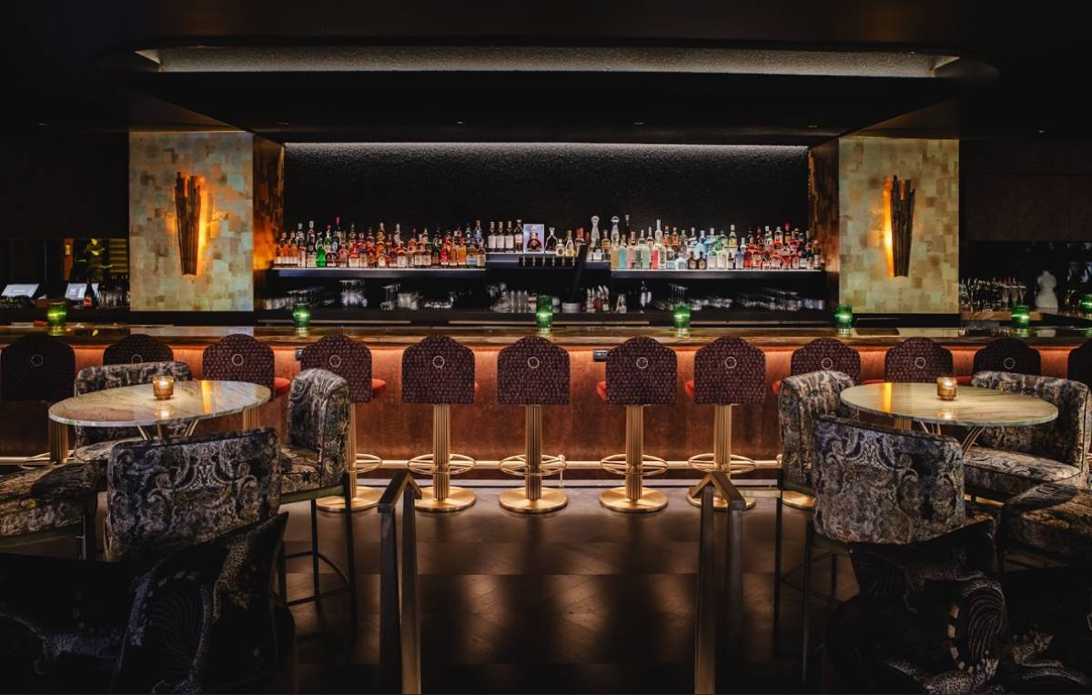 The interior of a fancy cocktail bar, with high-top tables and a row of chairs in front of a burnished bar