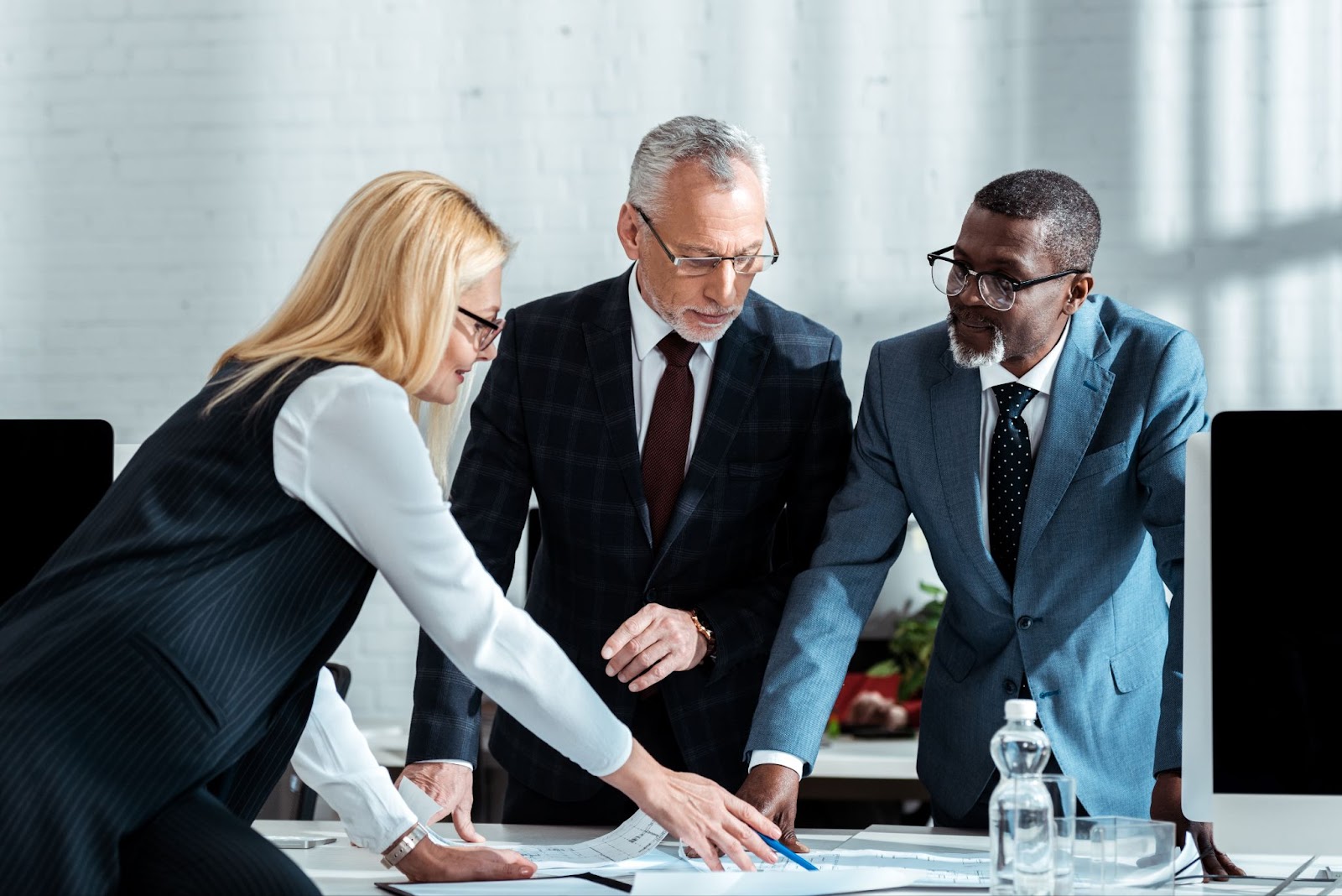 Three diverse business professionals discuss plans, with a woman pointing to documents laid out on the table.