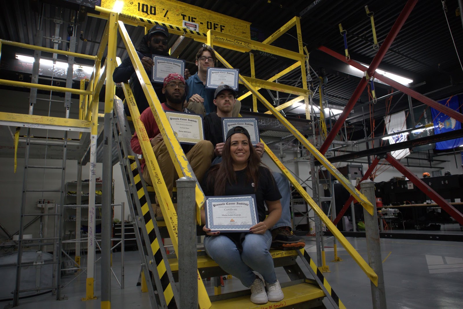 A group of five students posing on yellow industrial stairs, holding their Pinnacle Career Institute certificates. The background features safety signage and equipment, emphasizing a hands-on learning environment. The group looks proud and accomplished, representing diverse individuals celebrating their achievement.