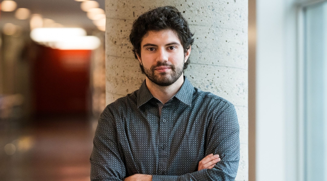 Dietitian with short curly hair and beard, wearing a patterned shirt, standing with arms crossed against a concrete pillar in a well-lit hallway.