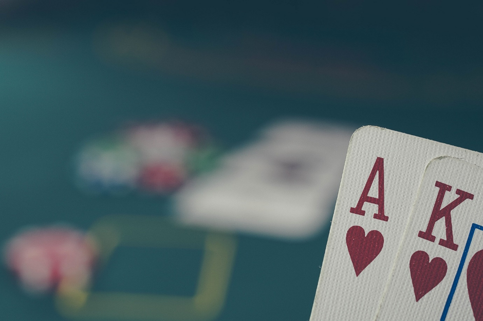 Close-up of Ace and King of hearts in a poker hand, with a blurred table and poker chips in the background.