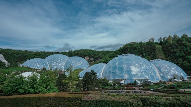 A series of large, clear geodesic domes set against lush green hills and a partly cloudy sky showcases innovative sustainable designs. The structures, surrounded by gardens and trees, beautifully blend nature with architecture, reflecting the visionary work of architects in community development.