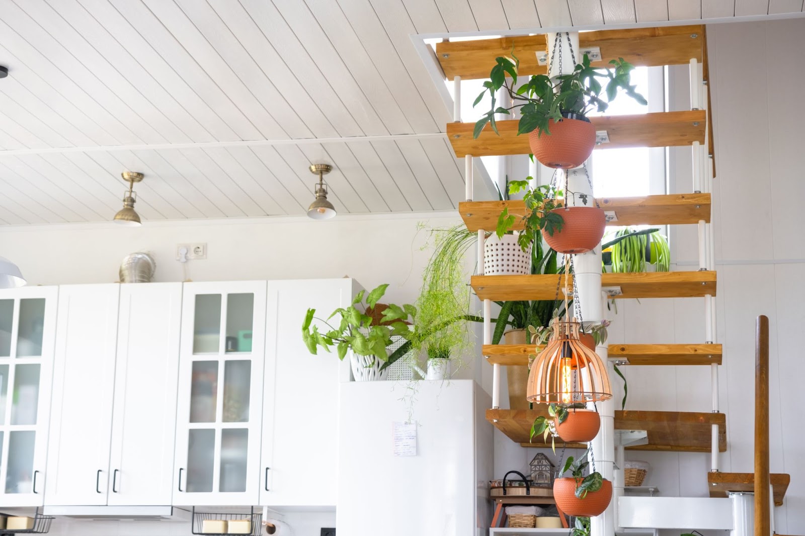 A modular metal staircase with wooden steps in a bright white interior with hanging plants beneath the stairs and white hanging cabinets.