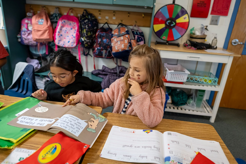 Two elementary age students reading together in a classroom. 
