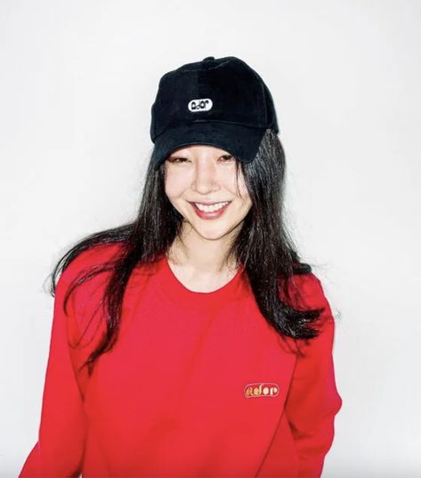 This contains an image of Min Hee Jin in a red shirt and black hat smiles at the camera while standing against a white wall