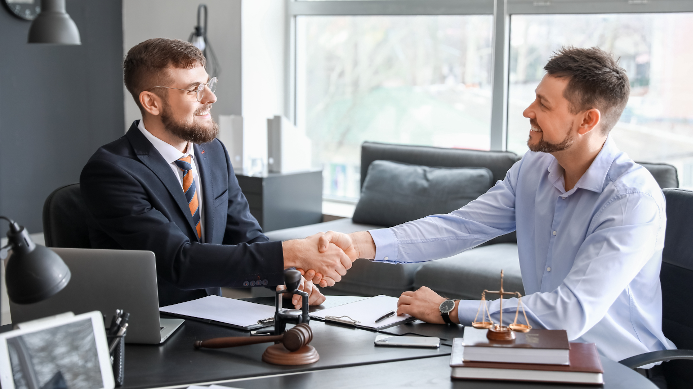 Two business professionals shaking hands in law office with legal scales and gavel on desk during contract negotiation