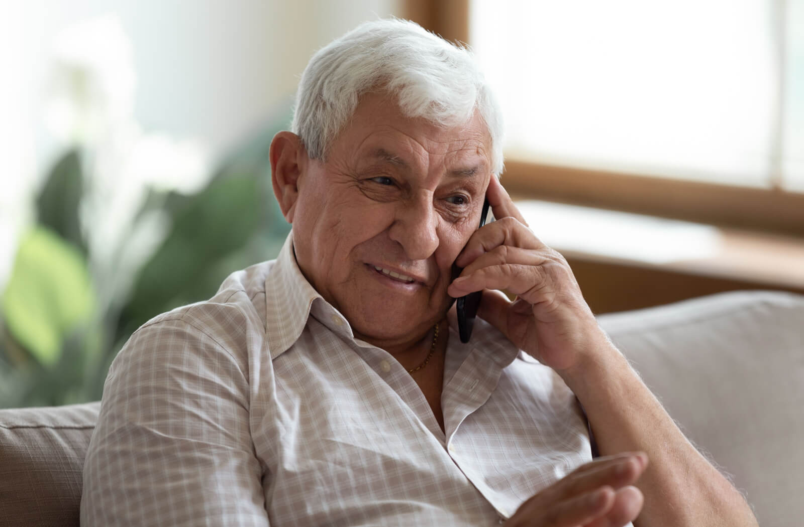 An older adult with dementia sitting on the couch and smiling while talking on the phone to someone.