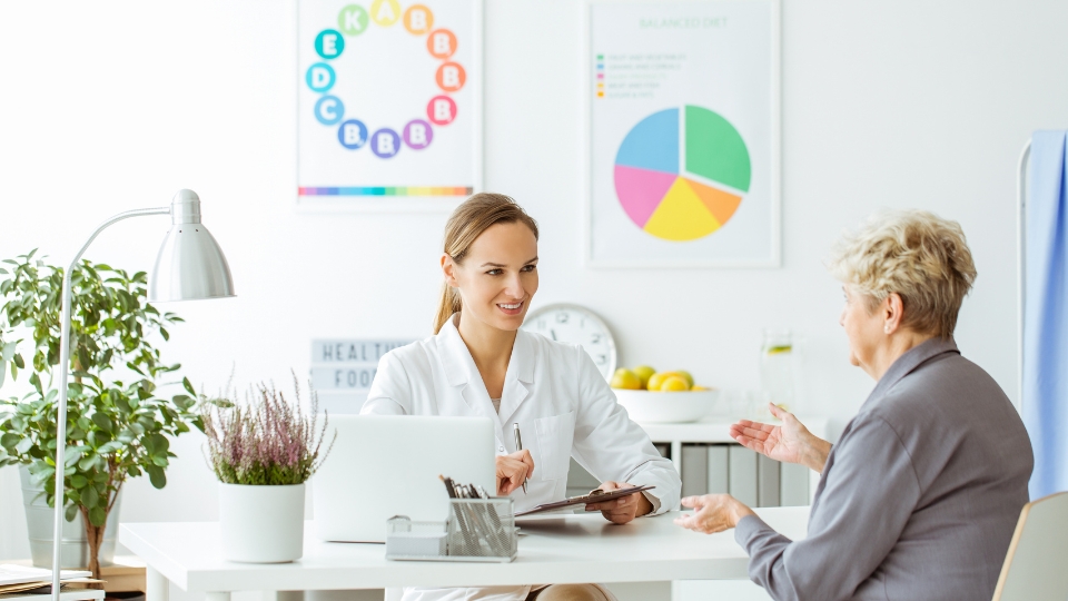 A smiling nutritionist dietitian talking with an elderly patient in a bright, modern office, with colourful posters illustrating nutritional concepts hanging on the wall in the backgrou