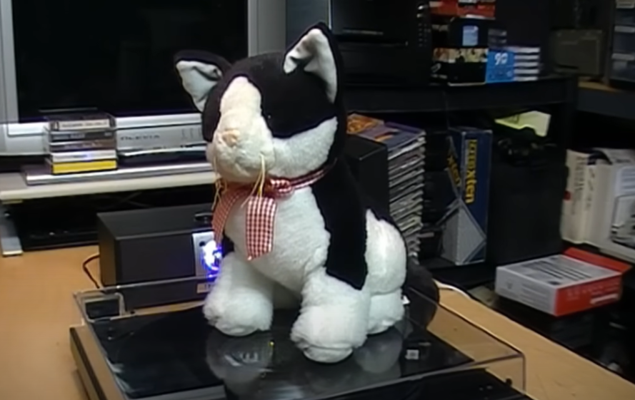 A stuffed black and white cat with a checkered ribbon sitting on top of a turntable in a room filled with electronic equipment.