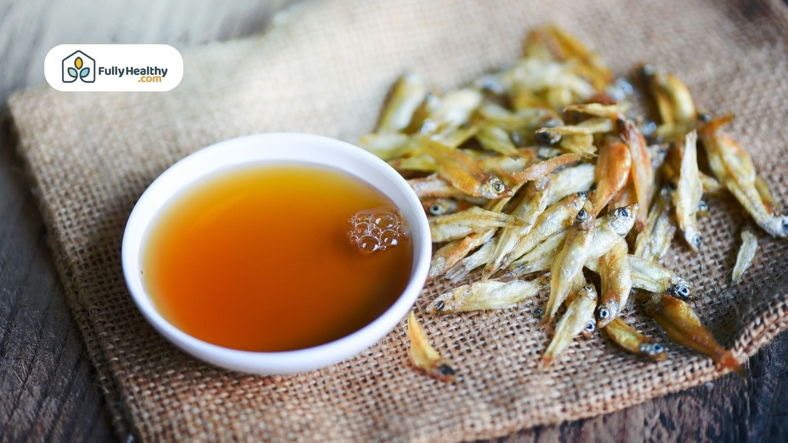 Fish sauce in white bowl beside dried anchovies on burlap