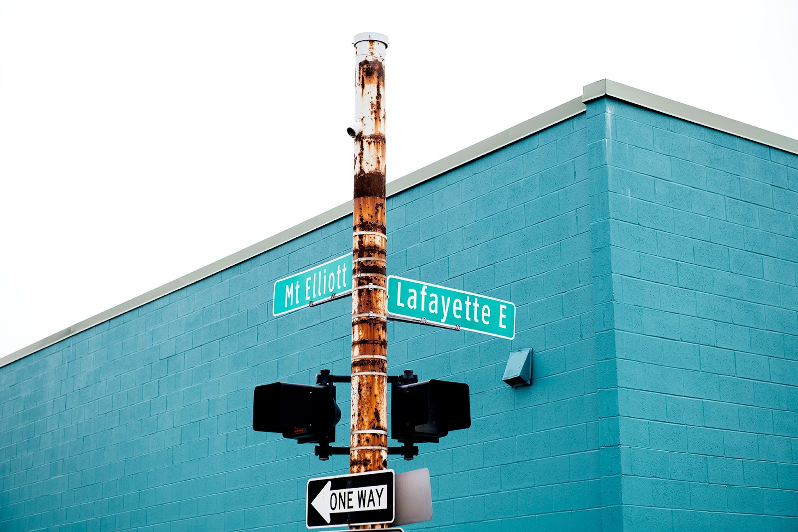 A street corner featuring a rusted pole with directional street signs for "Mt Elliott" and "Lafayette E," mounted above a traffic signal. The background showcases a vibrant teal-painted brick building under an overcast sky. A "One Way" sign is visible at the bottom, adding to the urban aesthetic of the scene.