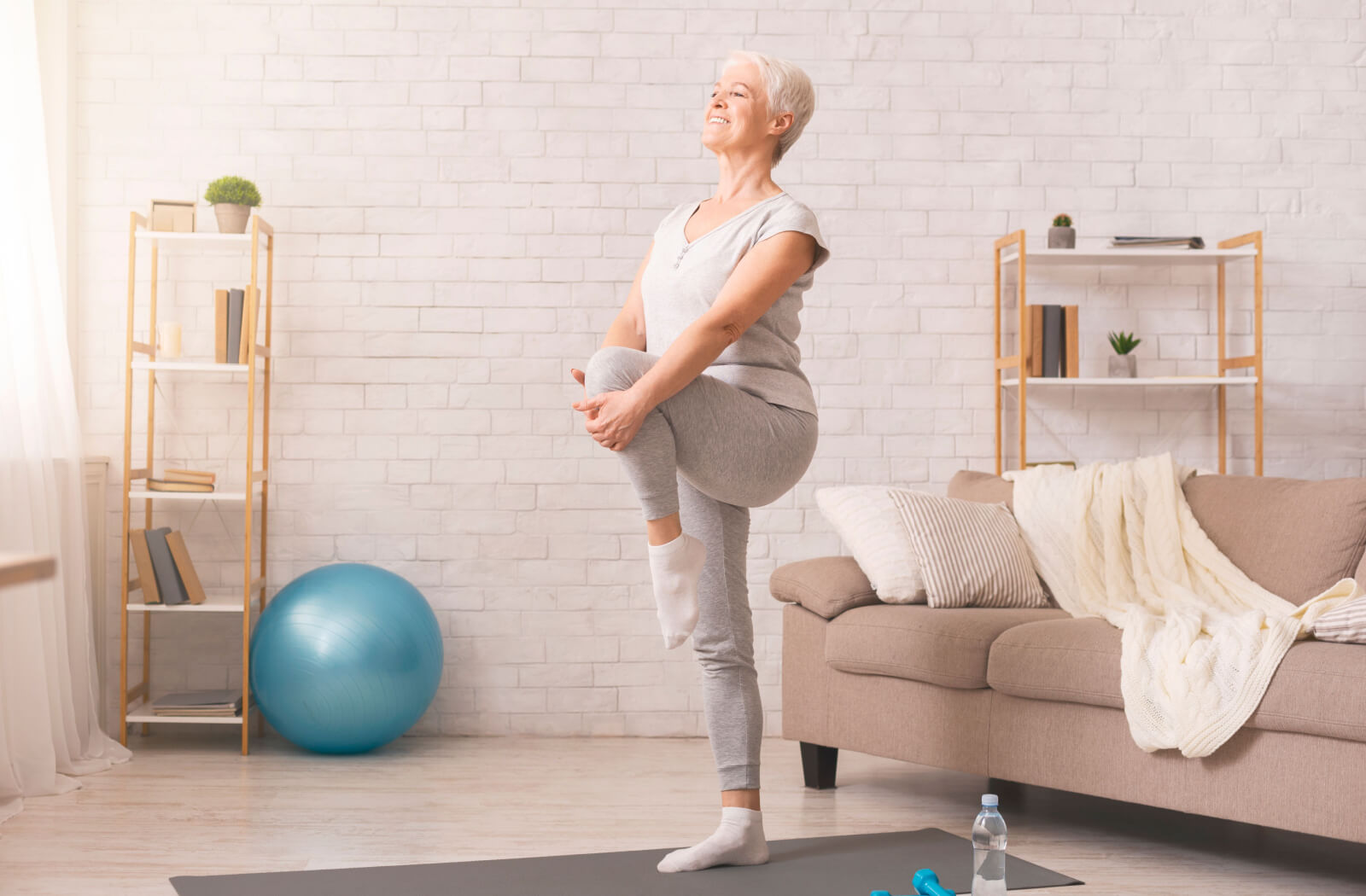 An older adult standing in a sunlit living room, holding their leg during a low-impact leg lift workout.
