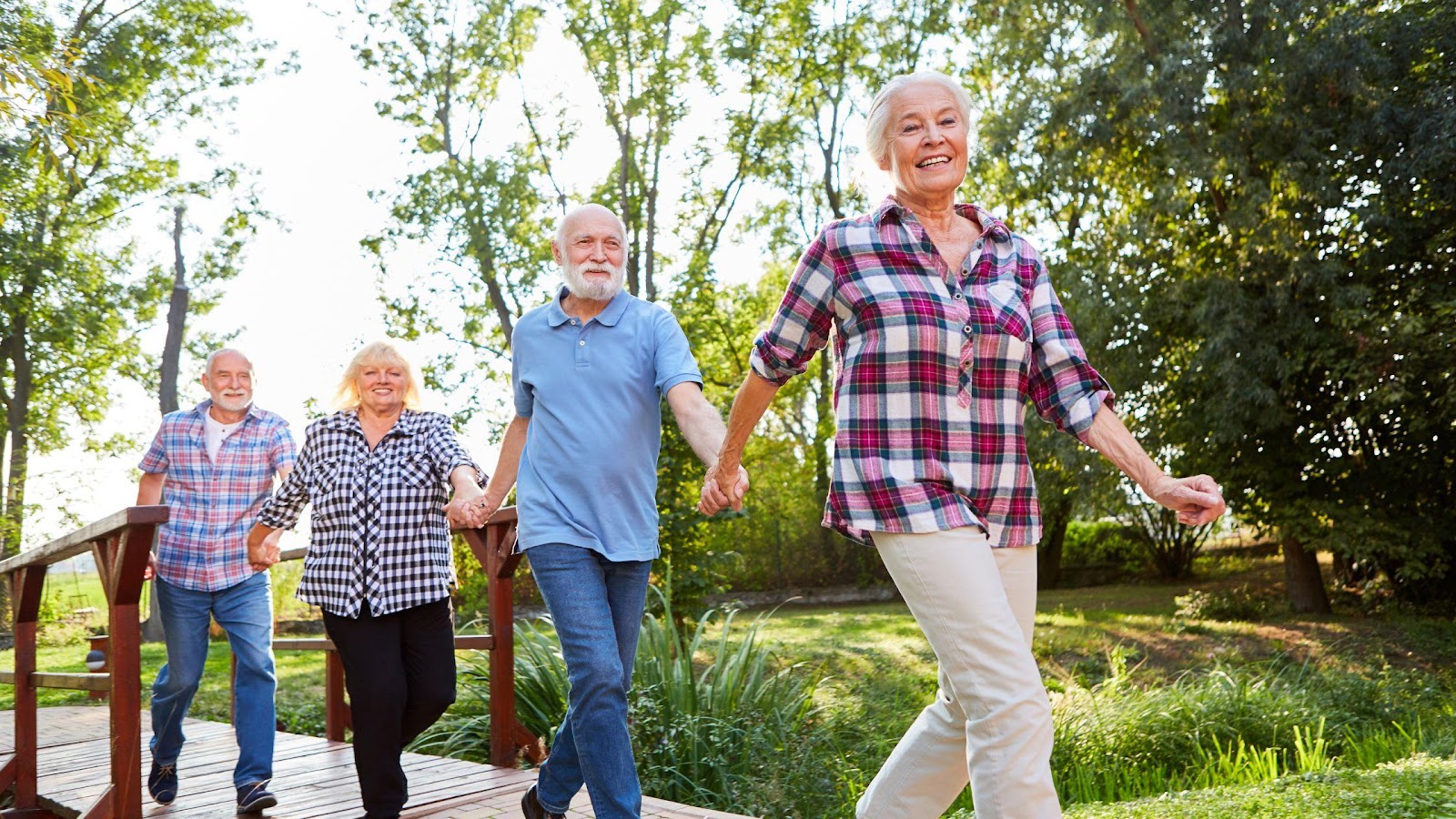 Four happy senior friends joyfully walking across a bridge in a lush park, embodying the ease and enjoyment of senior group travel excursions.