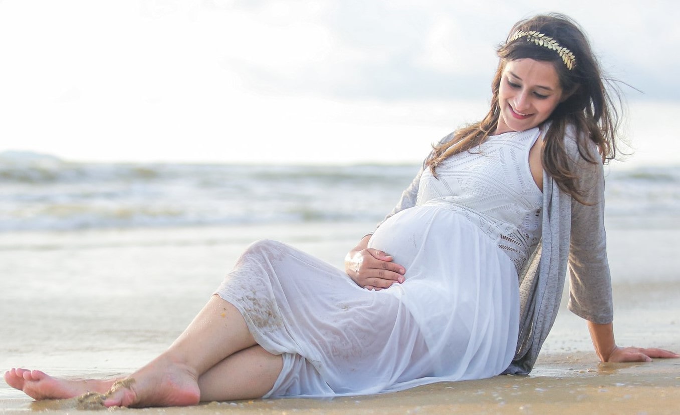 Maternity photoshoot by the beach: Expecting mom in a serene pose on the sand, wearing a white dress.