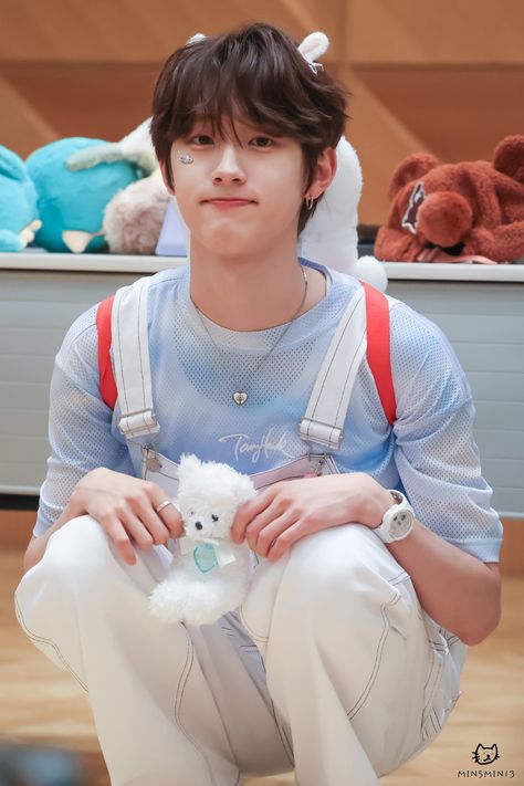 This contains an image of DRIPPIN Minseo's sitting on the floor holding a white teddy bear in his lap and wearing suspenders