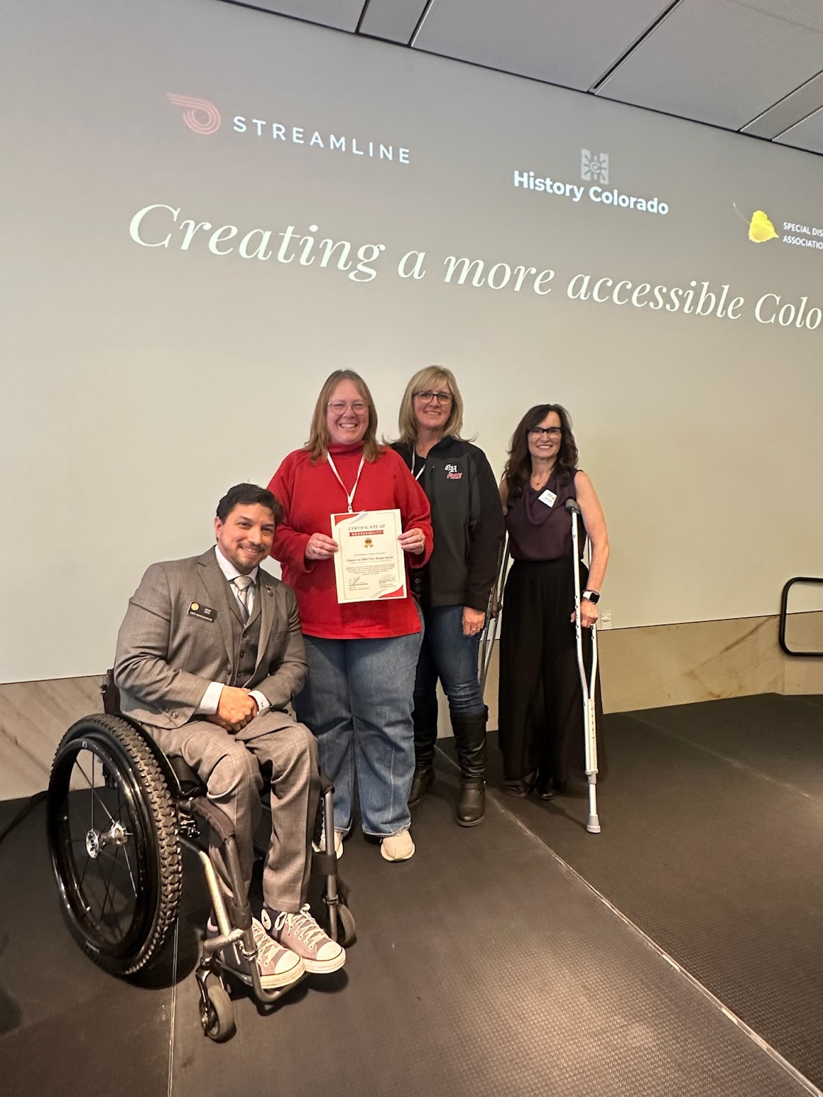 This image shows a group of four individuals standing on a stage in front of a projected screen with the text "Creating a more accessible Colorado" and logos from organizations like Streamline, History Colorado, and BCAI (Black Cube Artist Incubator). On the left, a man in a wheelchair wearing a grey suit smiles while sitting. Next to him, a woman in a red shirt holds a certificate, smiling. Two other women stand beside her: one wearing a dark jacket, and the other with crutches, dressed in black, smiling as well. 