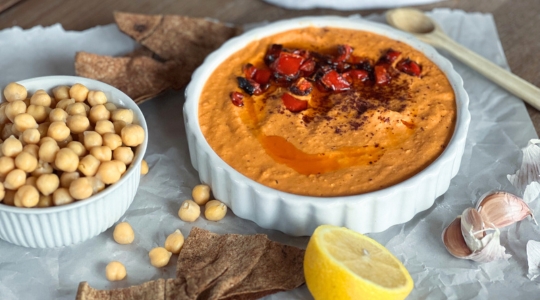 A bowl of red pepper hummus garnished with roasted red pepper pieces, served alongside chickpeas, pita chips, a lemon wedge, and garlic cloves.