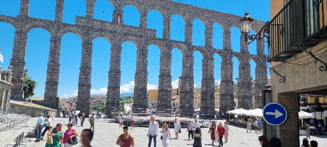 A group of people standing in front of a large stone structure with Aqueduct of Segovia in the backgroundDescription automatically generated