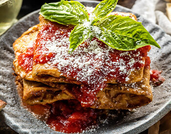 A serving of golden-brown fried lasagna topped with marinara sauce, fresh basil, and dusted with grated cheese, plated on a dark ceramic dish.