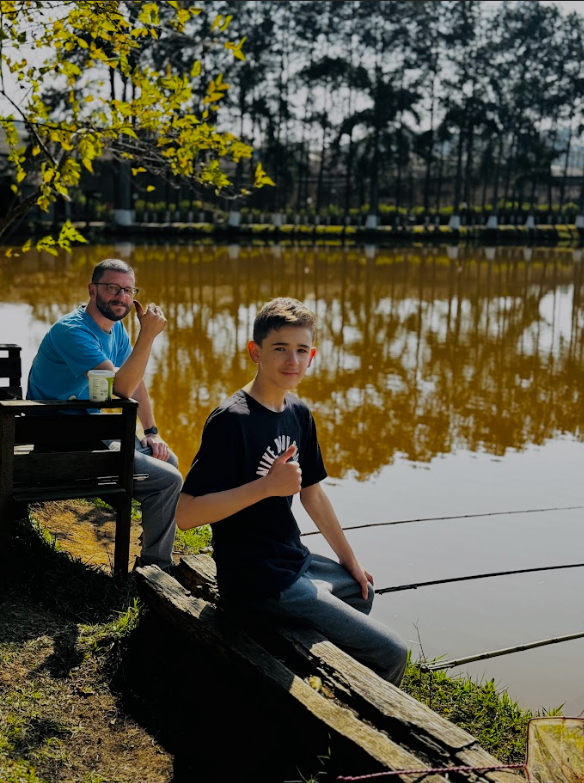 Pai e filho pescando à beira de um lago em um dia ensolarado, com árvores ao fundo e um ambiente tranquilo, ideal para momentos de lazer e conexão com a natureza.