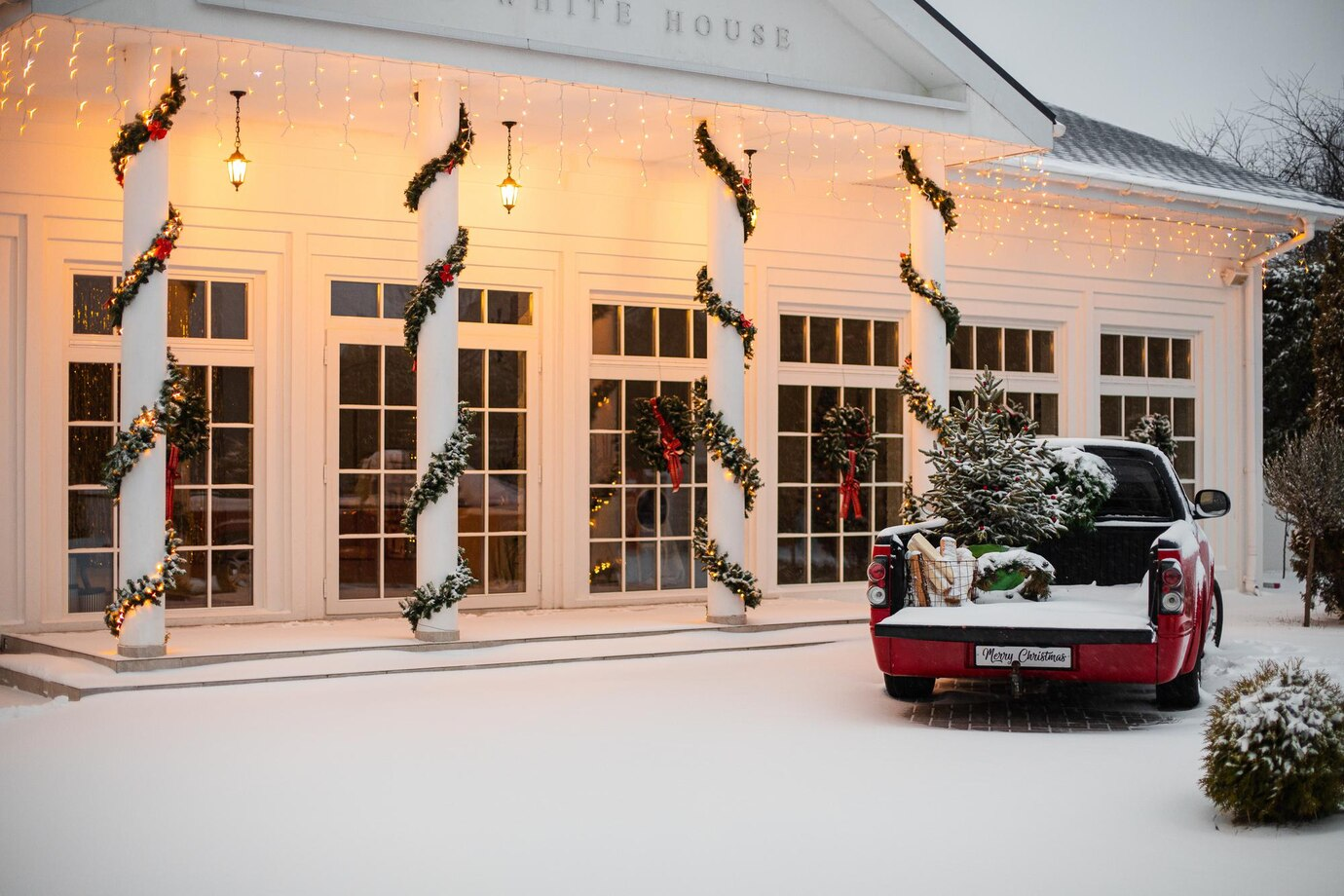 A festive scene featuring a red pickup truck loaded with snow-covered Christmas trees and decorations, parked in front of a brightly lit building with columns wrapped in garlands and string lights, surrounded by a snowy landscape