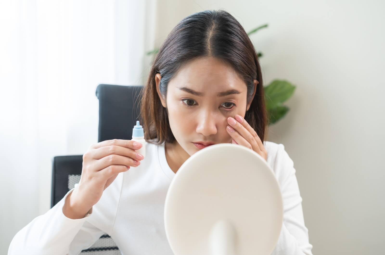 A young woman sitting with a bottle of eye drops for dry eye in her right hand and looking at a desk mirror while holding her left eye open with her left hand.