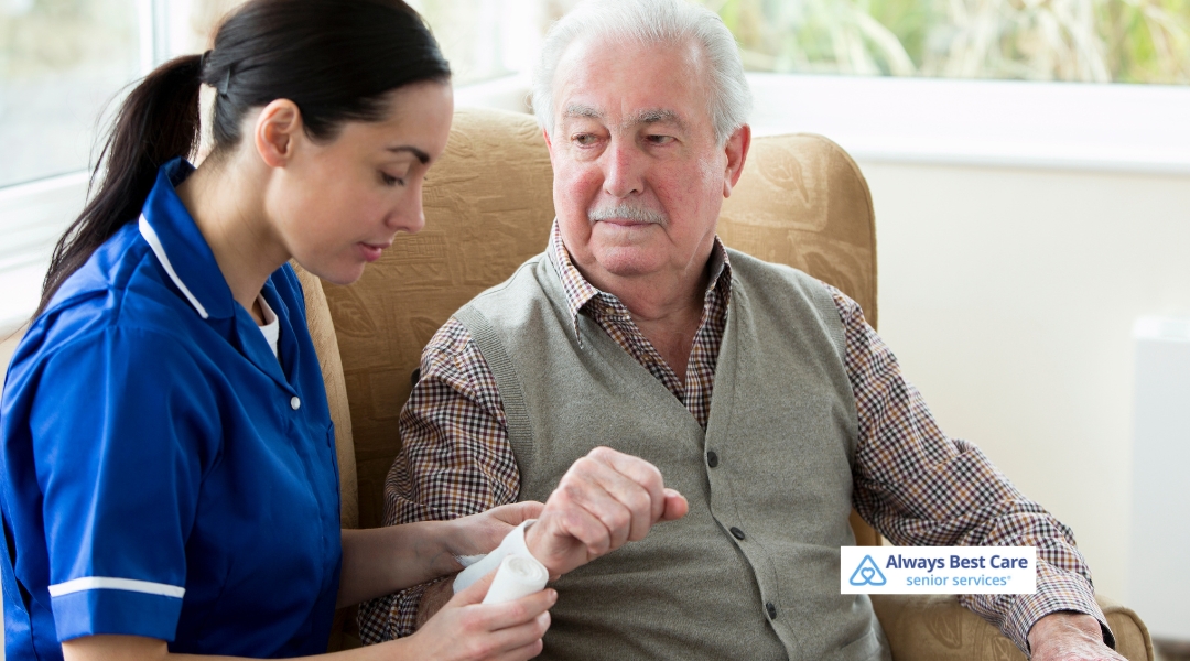  Female caregiver gently wrapping the wrist of an elderly man seated in an armchair, showcasing attentive in-home care for seniors.

