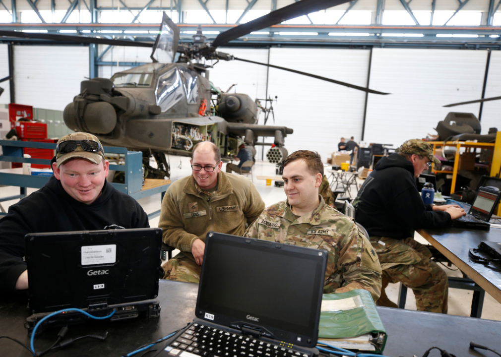 Military personnel on computers in front of a helicopter in a bright warehouse.