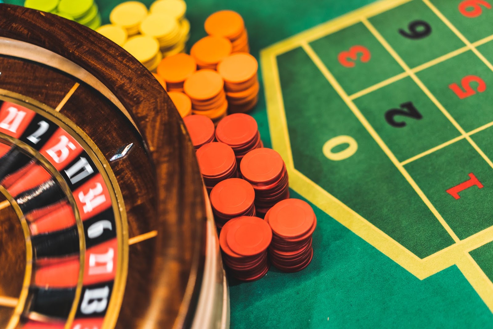 A large plan of a roulette wheel with a wooden edge and red and black numbers surrounded by piles of colored chips on a green roulette table with visible numbers at stake