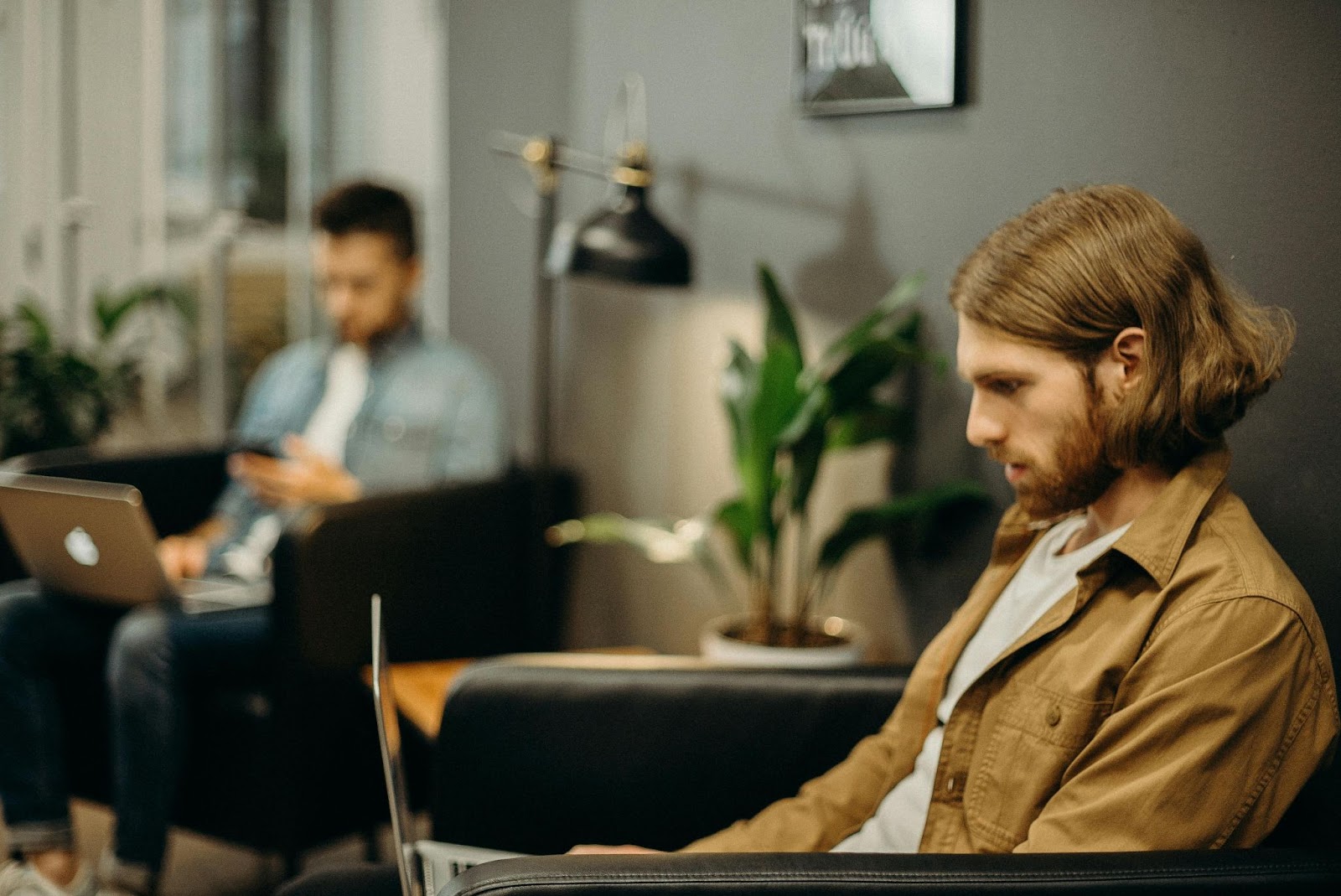 Two men are working on laptops in a modern, indoor setting. One man in the foreground is focused on his screen, while another in the background is using his phone. There are plants and a lamp in the room.