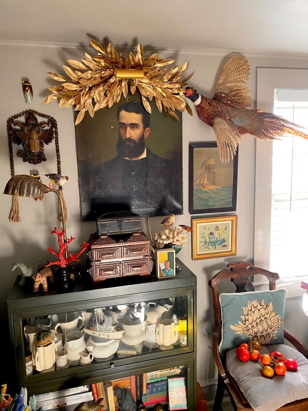 A corner of the room features a dark portrait of a beaded man on the wall, topped with a decorative golden laurel wreath. A taxidermy pheasant is mounted beside the portrait. Below, a glass-fronted cabinet displays a collection of white ceramics and pottery. The cabinet top is adorned with various decorative objects, including a red coral sculpture and a woven box. A wooden chair with a seashell[-themed cushion holds a few ripe tomatoes is placed next to the cabinet.